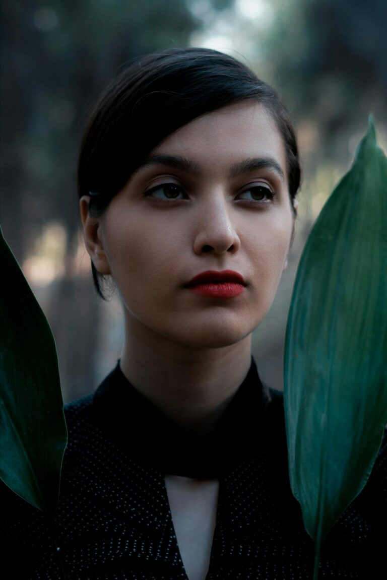 Young dreamy female in black clothes standing with verdant leaves on blurred woodland background and looking away